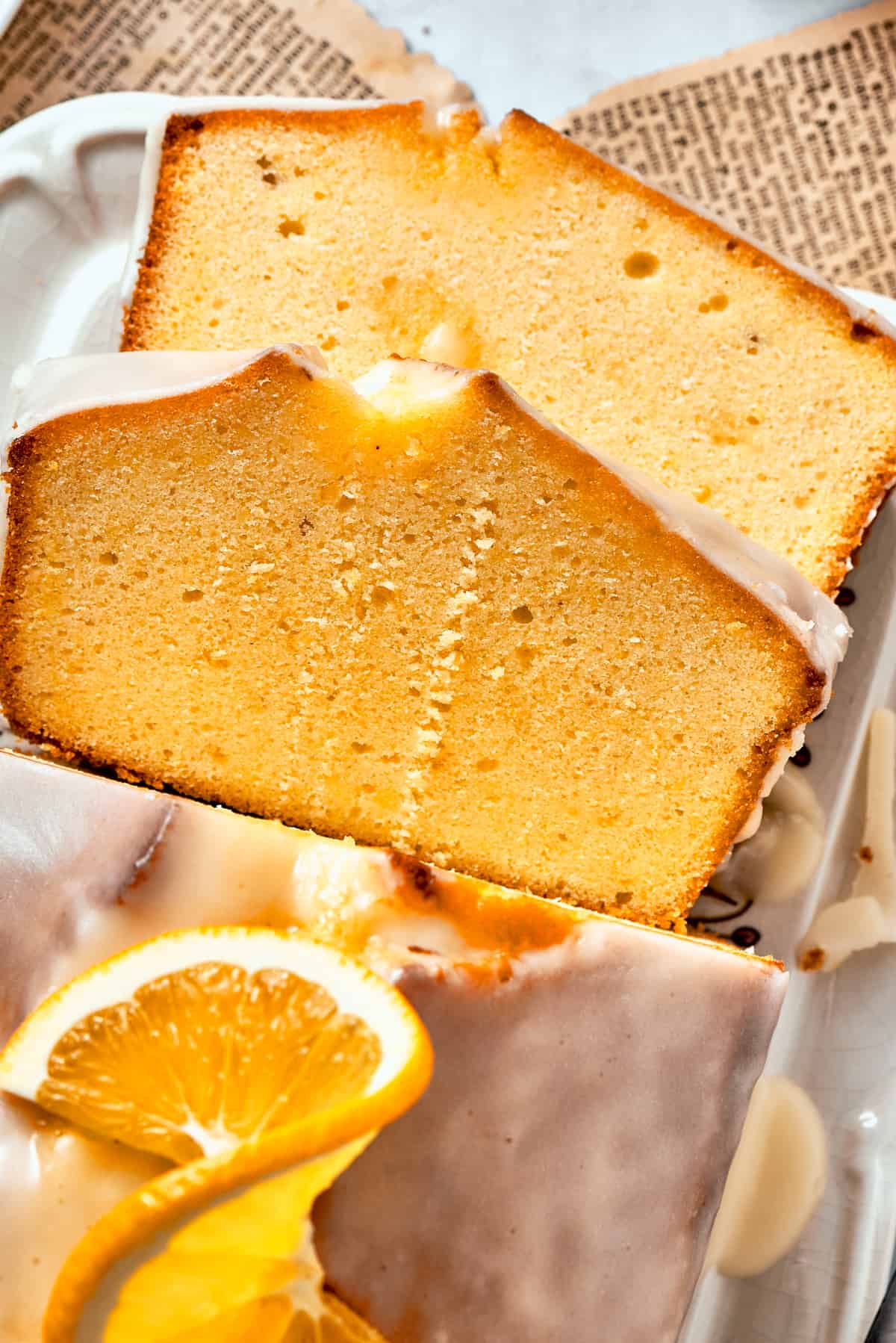 Two slices of glazed orange cake rest atop each other, with the rest of the loaf visible nearby topped with orange slices.