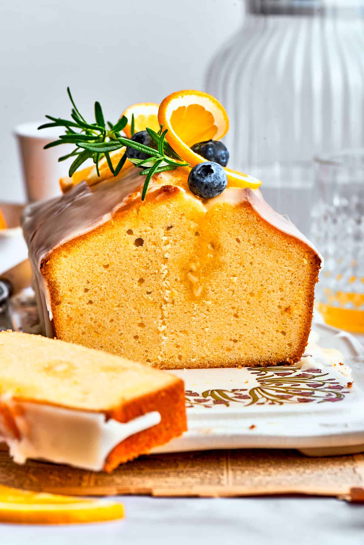 A side view of a slice of orange cake and the entire loaf, topped with blueberries, fresh rosemary, and orange slices.