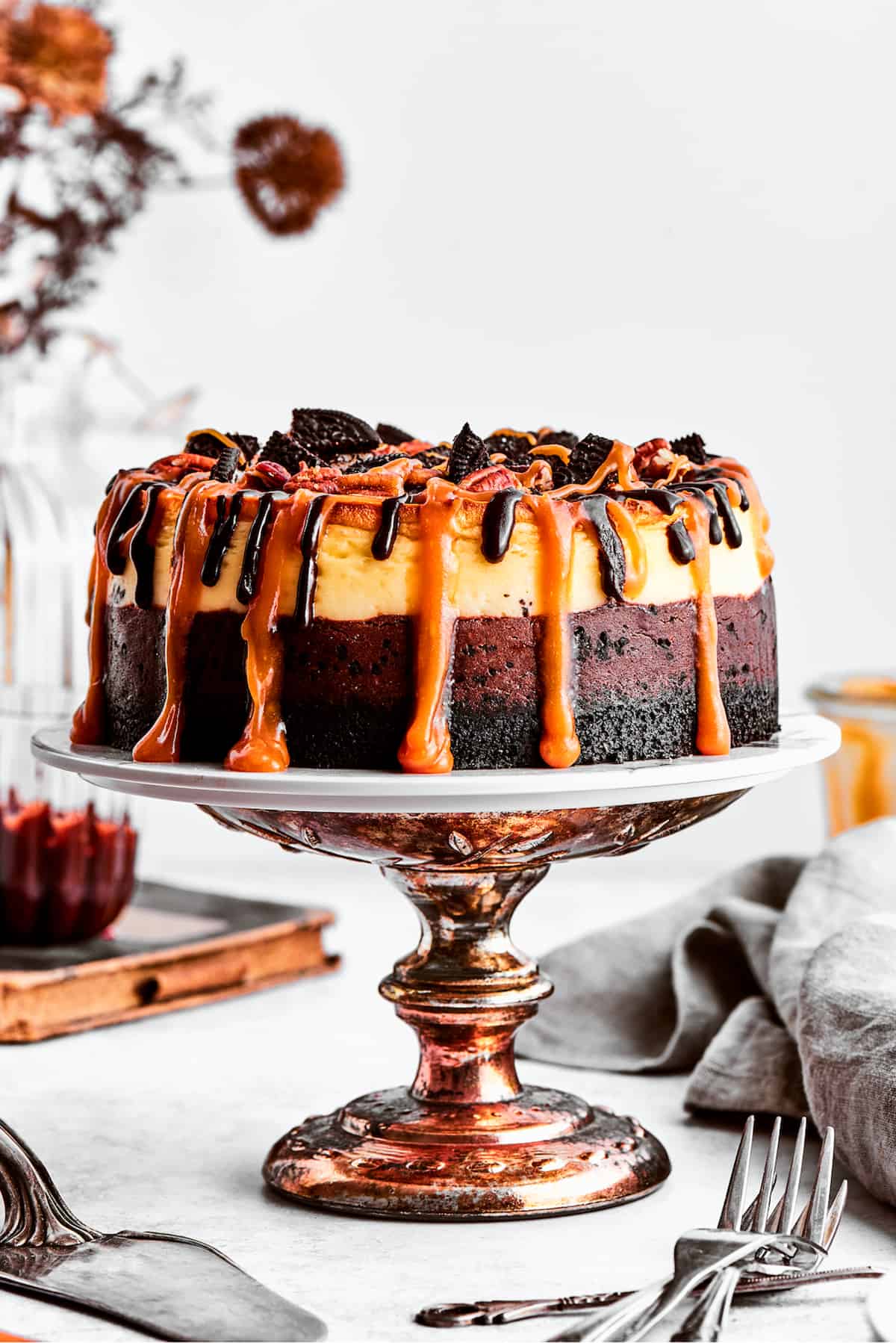 A turtle cheesecake dripping with caramel sauce is displayed on a bronze cake stand.