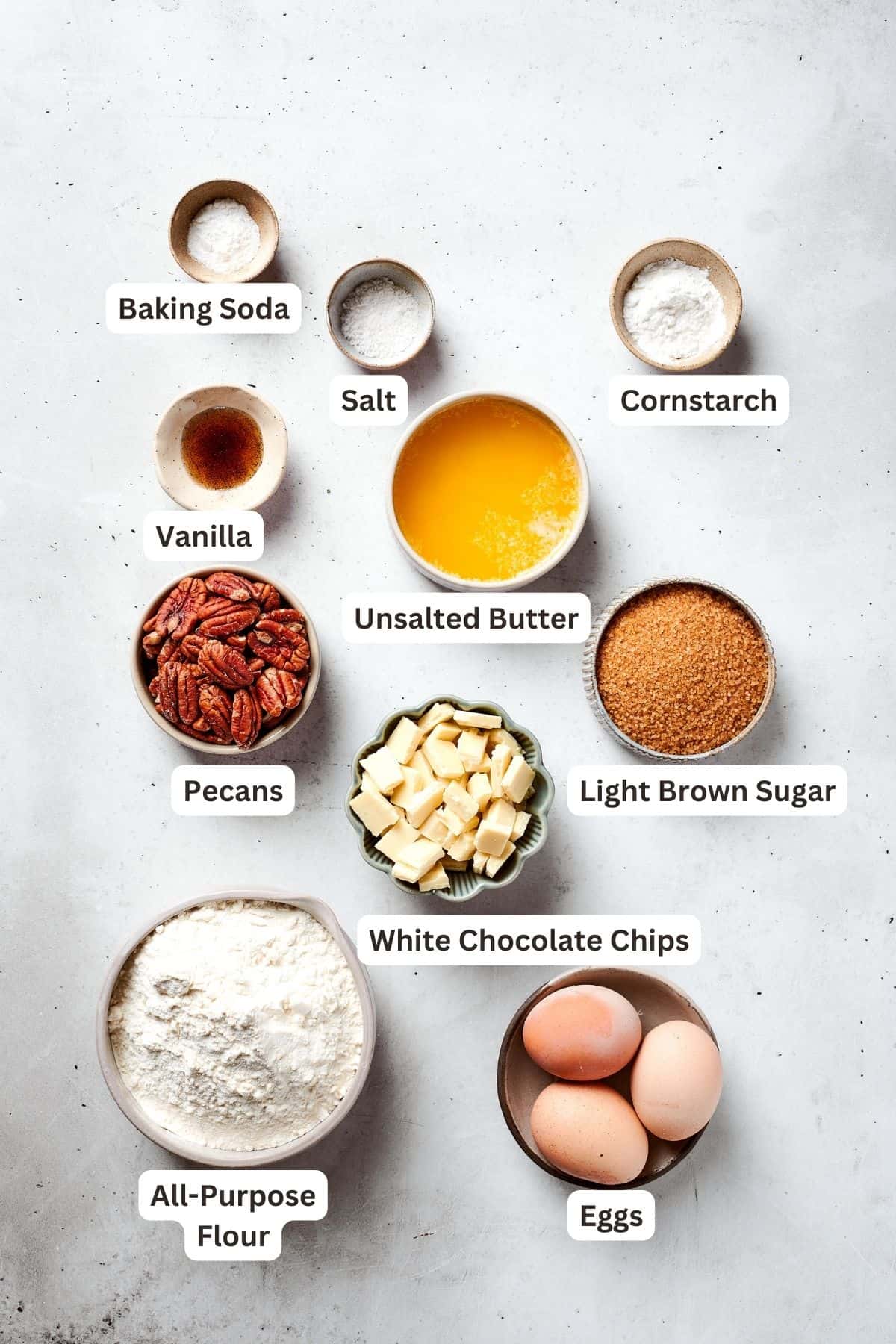The ingredients for blonde brownies are shown portioned out: flour, cornstarch, baking soda, salt, vanilla, brown sugar, melted butter, eggs, pecans, white chocolate chips.