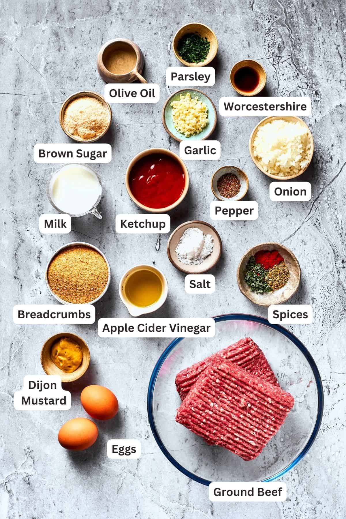 The ingredients for brown sugar meatloaf are shown portioned out: meat, ketchup, brown sugar, apple cider vinegar, salt and pepper, herbs, onion, garlic, breadcrumbs, Worcestershire sauce, eggs, olive oil.