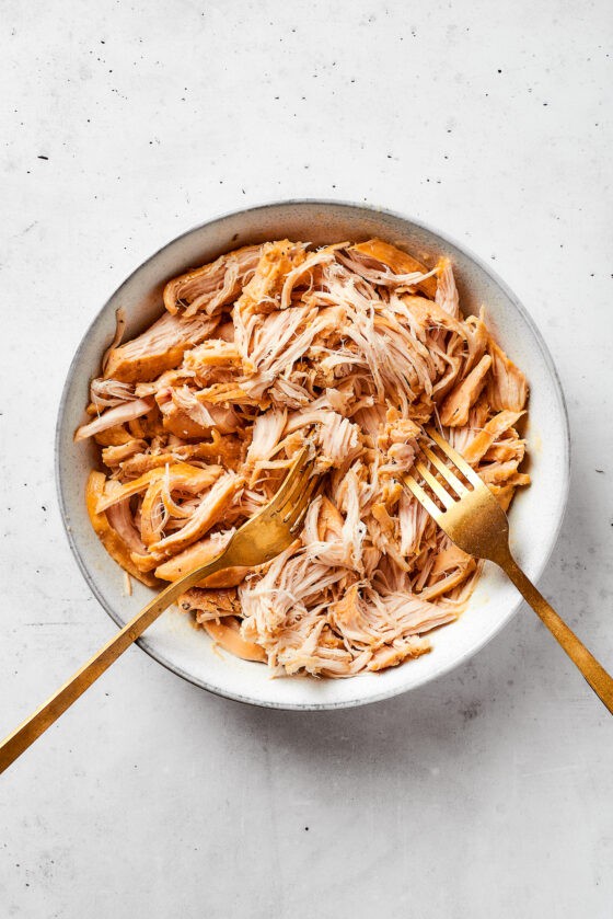 Two forks work to shred chicken in a white bowl.