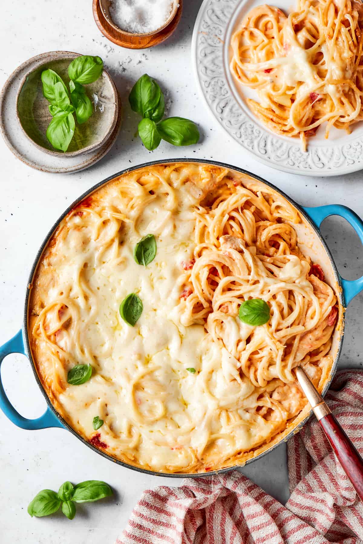 A pot of cheesy chicken spaghetti on a table with a bowl of fresh basil leaves next to it.