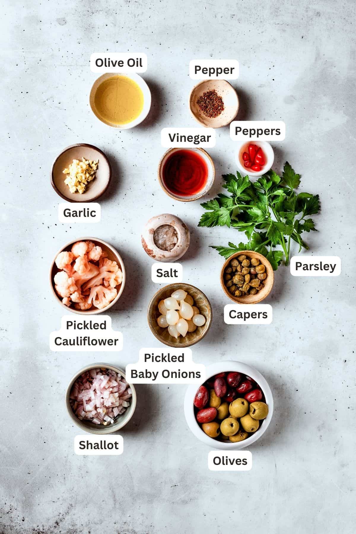 The ingredients for olive salad are shown: olives, peppers, pickled cauliflower, vinegar, parsley, capers, pickled baby onions, garlic, olive oil.