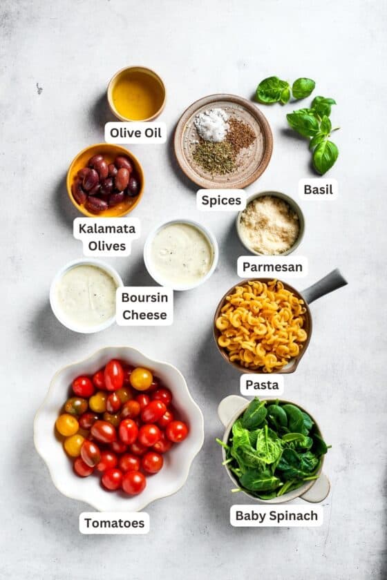 Ingredients needed to make Boursin pasta are shown: Boursin cheese, cherry tomatoes, grape tomatoes, Parmesan, Kalamata olives, pasta, baby spinach, basil, Italian seasoning, olive oil, salt and pepper.