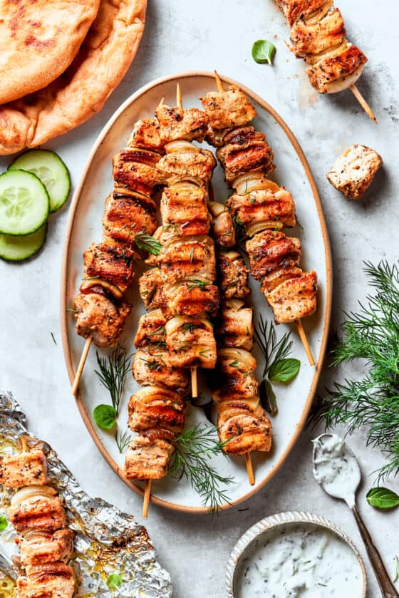 A platter of grilled chicken souvlaki on skewers surrounded by herbs and cucumber slices.
