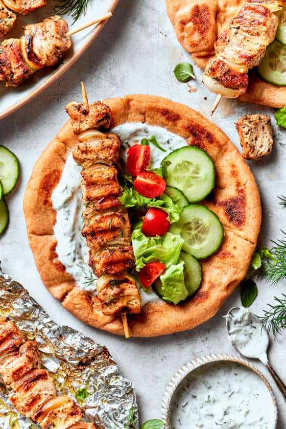 A skewer of chicken souvlaki is placed on a piece of flatbread alongside sliced cucumbers and tomatoes.