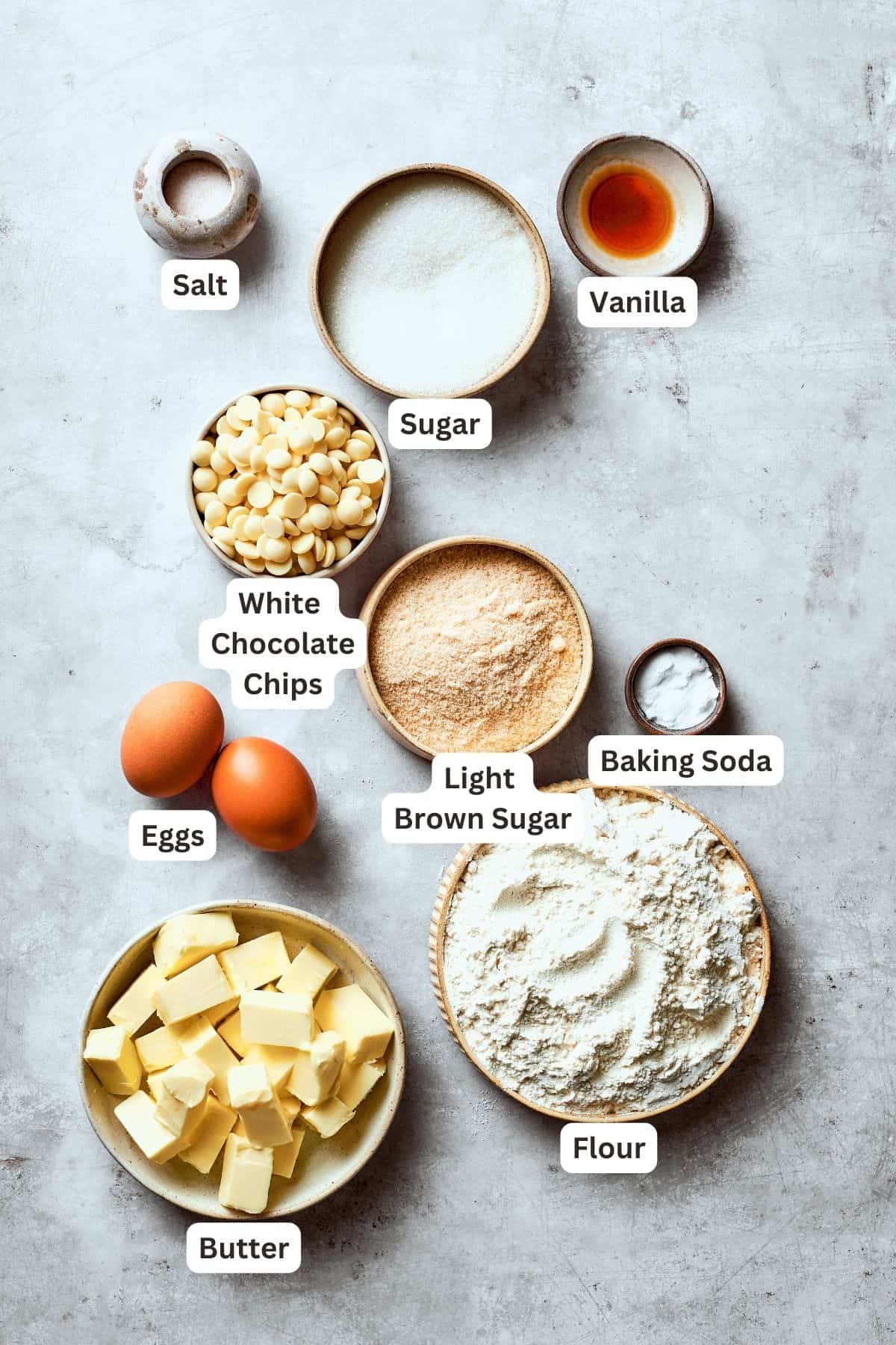 Ingredients for white chocolate chip cookies are shown labelled and portioned out: white chocolate chips, flour, butter, granulated sugar, brown sugar, baking soda, eggs, vanilla, salt.