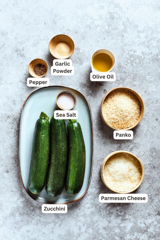 Ingredients for zucchini chips are shown labeled and portioned out: zucchini, garlic powder, pepper, olive oil, panko, salt, parmesan cheese.