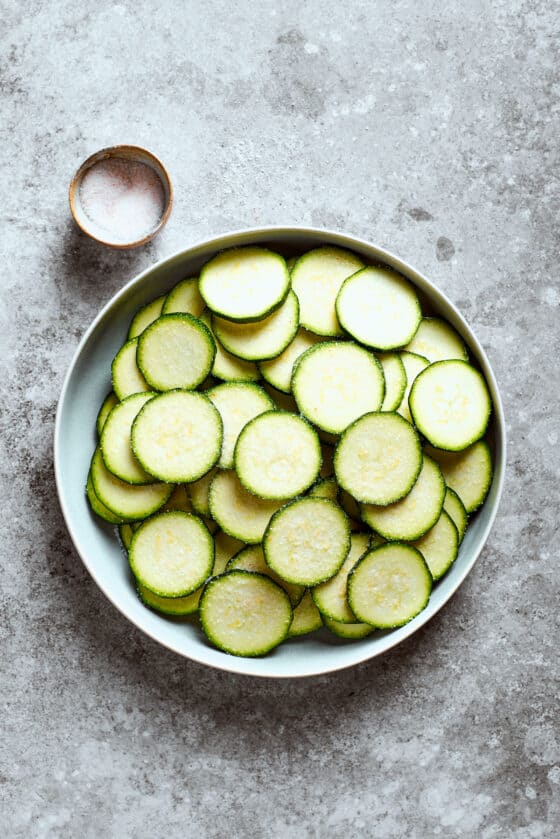 A bowl of sliced zucchini next to a small bowl of salt.