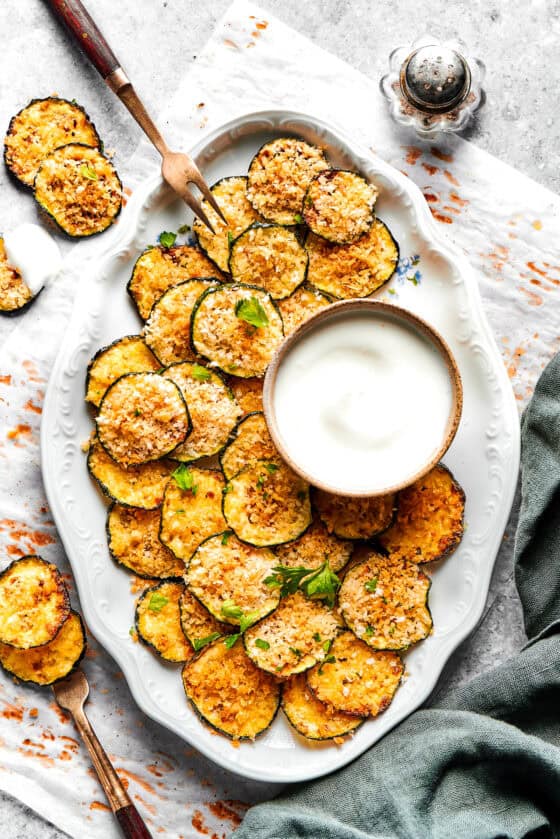 Baked zucchini chips served on an oval white platter with a bowl of dip.