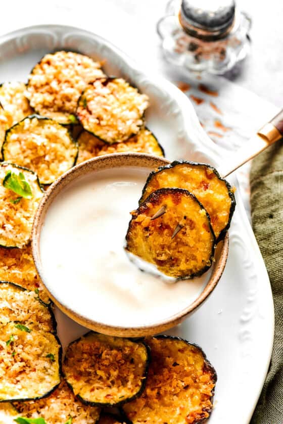 Two crispy zucchini chips in a bowl of yogurt on a platter with more chips.