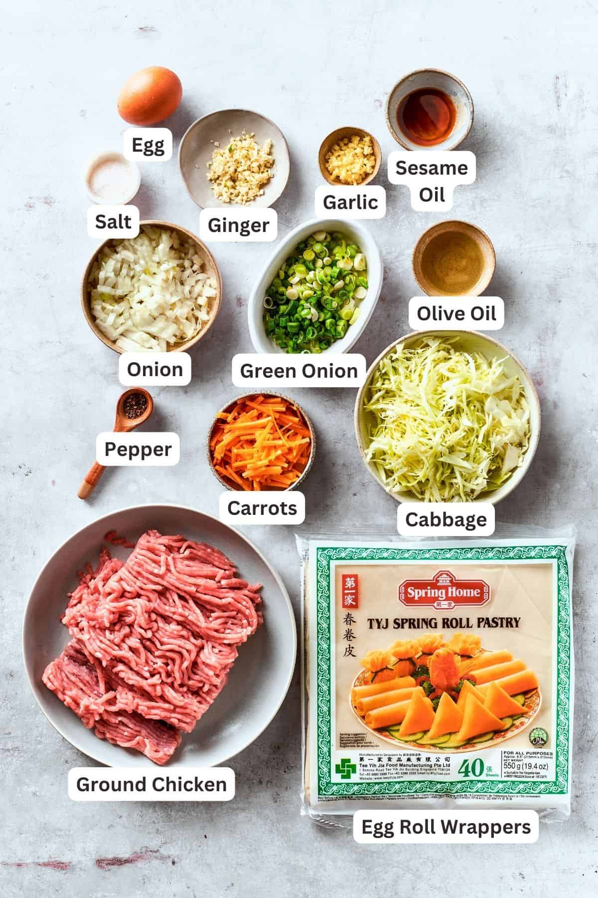 The ingredients for chicken egg rolls are shown portioned out and labelled: ground chicken, shredded carrots and cabbage, onion, garlic, olive oil ginger, sesame oil, garlic, green onions, egg roll wrappers, salt and pepper.