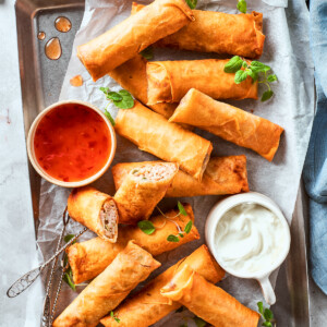 A bowl of red sauce and a bowl of white sauce are served on a serving tray full of chicken egg rolls.