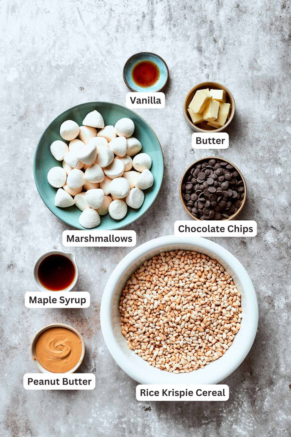 Ingredients for peanut butter rice crispy treats are portioned and labelled: mini marshmallows, chocolate chips, butter, maple syrup, peanut butter vanilla, rice cereal.