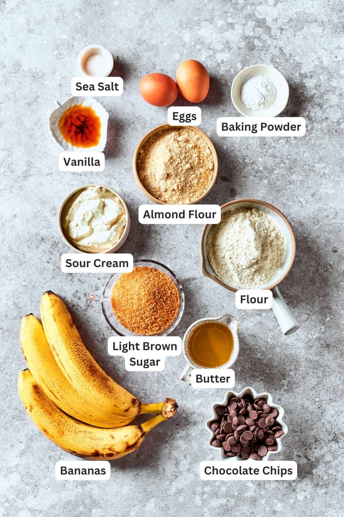 Ingredients for sour cream banana bread are labelled and portioned out: bananas, brown sugar, almond flour, sour cream, all-purpose flour, baking powder, eggs, vanilla, salt, chocolate chips, butter.