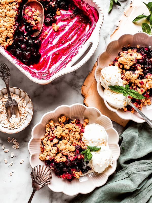Overhead image of two dessert bowls of blueberry crisp served with scoops of ice cream. There is a baking dish with the crisp placed near the bowls.