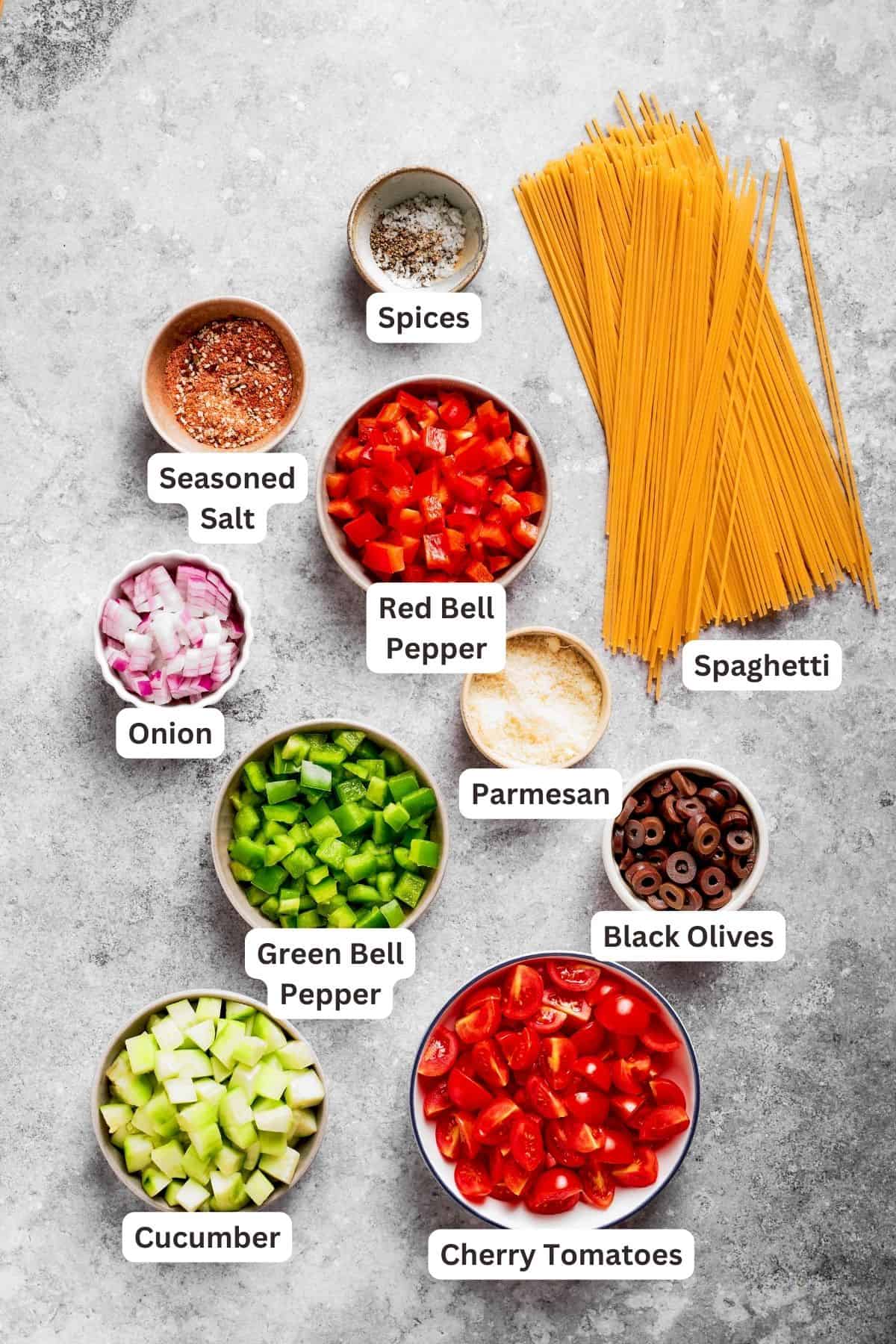 Ingredients for spaghetti salad are labeled and portioned out: spaghetti, cucumber, tomatoes, onion, bell peppers, black olives, parmesan.