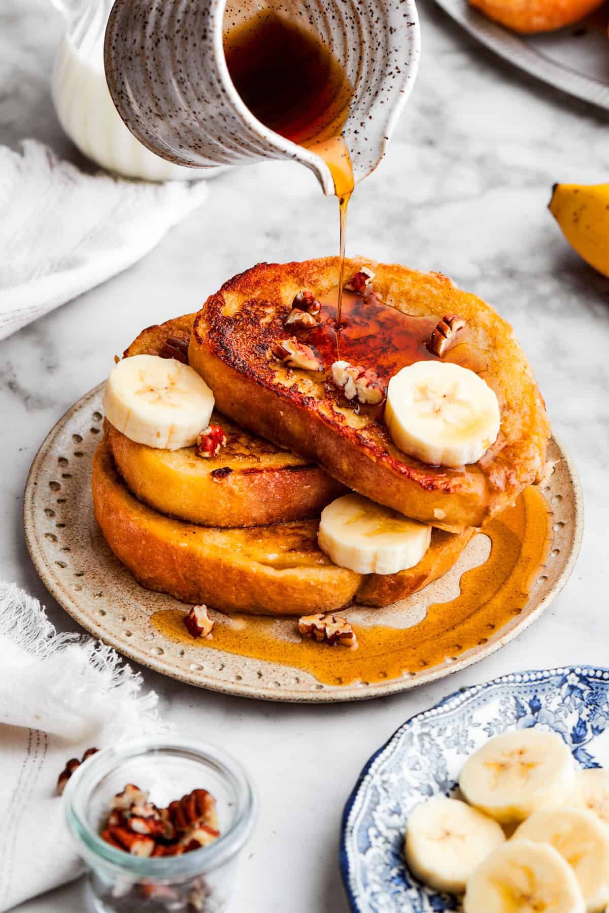 A pitcher pours maple syrup over a stack of banana French toast.