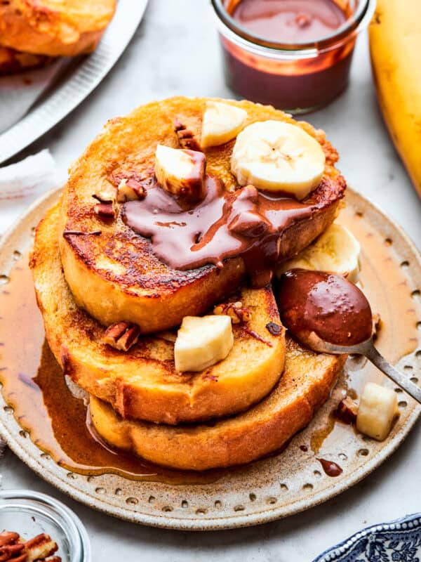 Three slices of French toast are stacked on top of each other on a plate and topped with banana slices and Nutella.