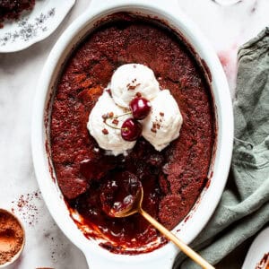 Chocolate cake in a casserole dish garnished with vanilla ice cream scoops, two cherries, with a gold-colored spoon set inside the cake.