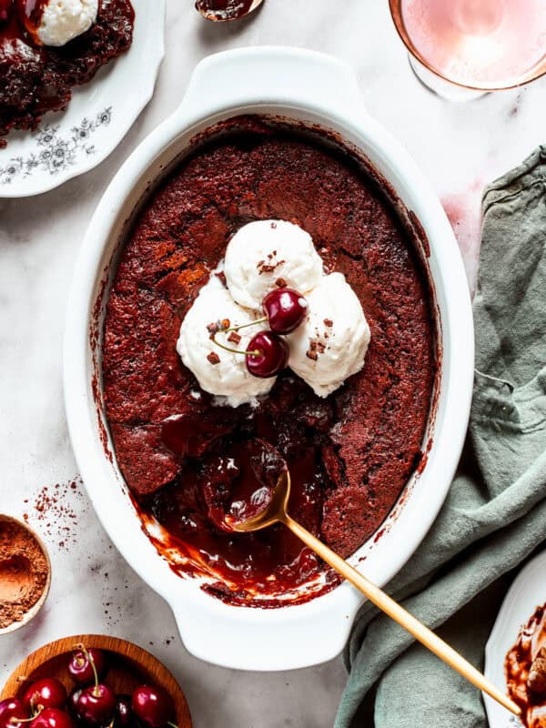 Chocolate cake in a casserole dish garnished with vanilla ice cream scoops, two cherries, with a gold-colored spoon set inside the cake.