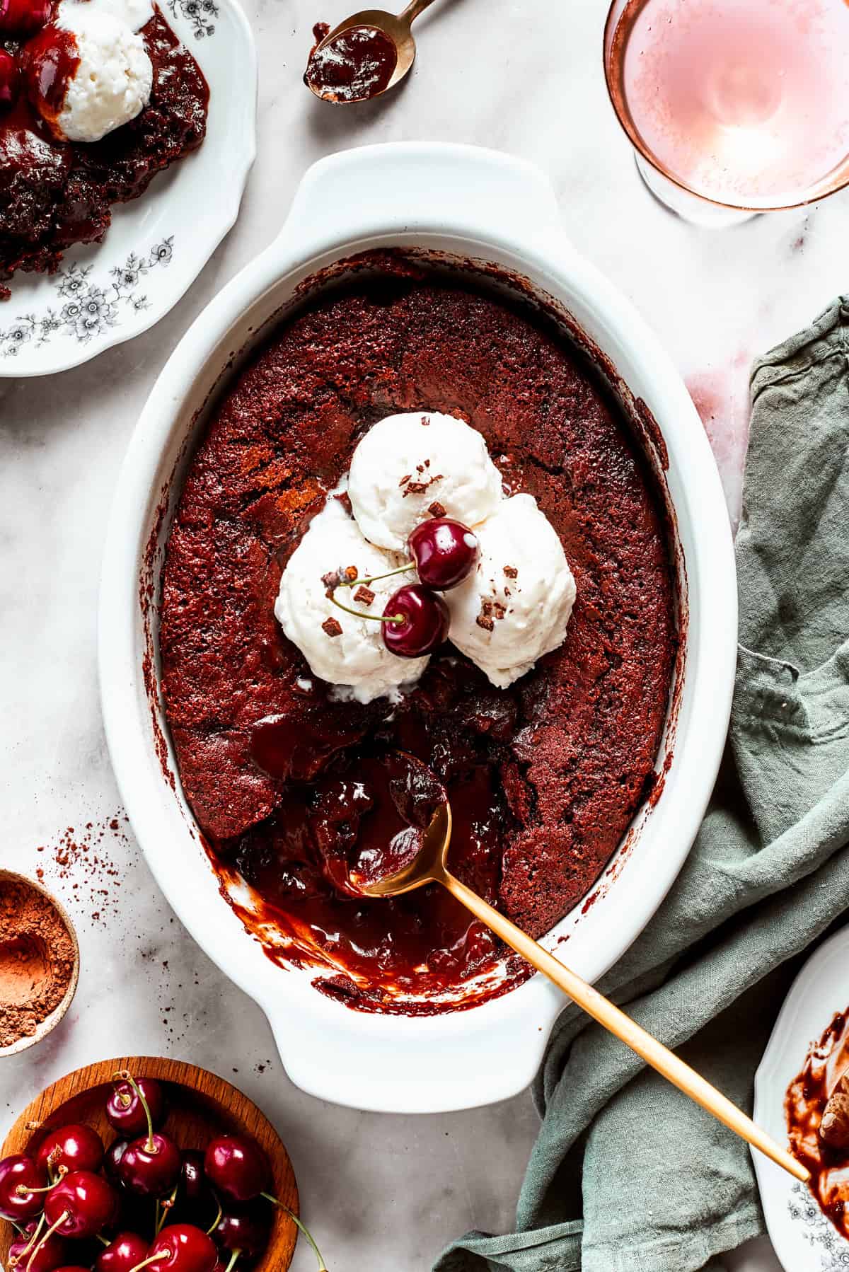 Chocolate cake in a casserole dish garnished with vanilla ice cream scoops, two cherries, with a gold-colored spoon set inside the cake.