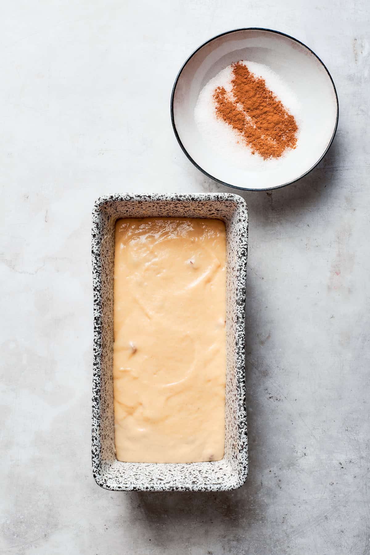 Amish friendship bread batter in a loaf pan with cinnamon sugar next to it.