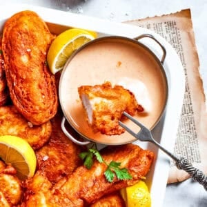 A piece of beer battered chicken is dipped into spicy sauce with more chicken pieces around it.