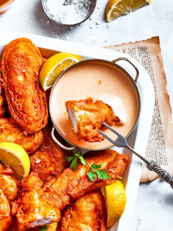 A piece of beer battered chicken is dipped into spicy sauce with more chicken pieces around it.