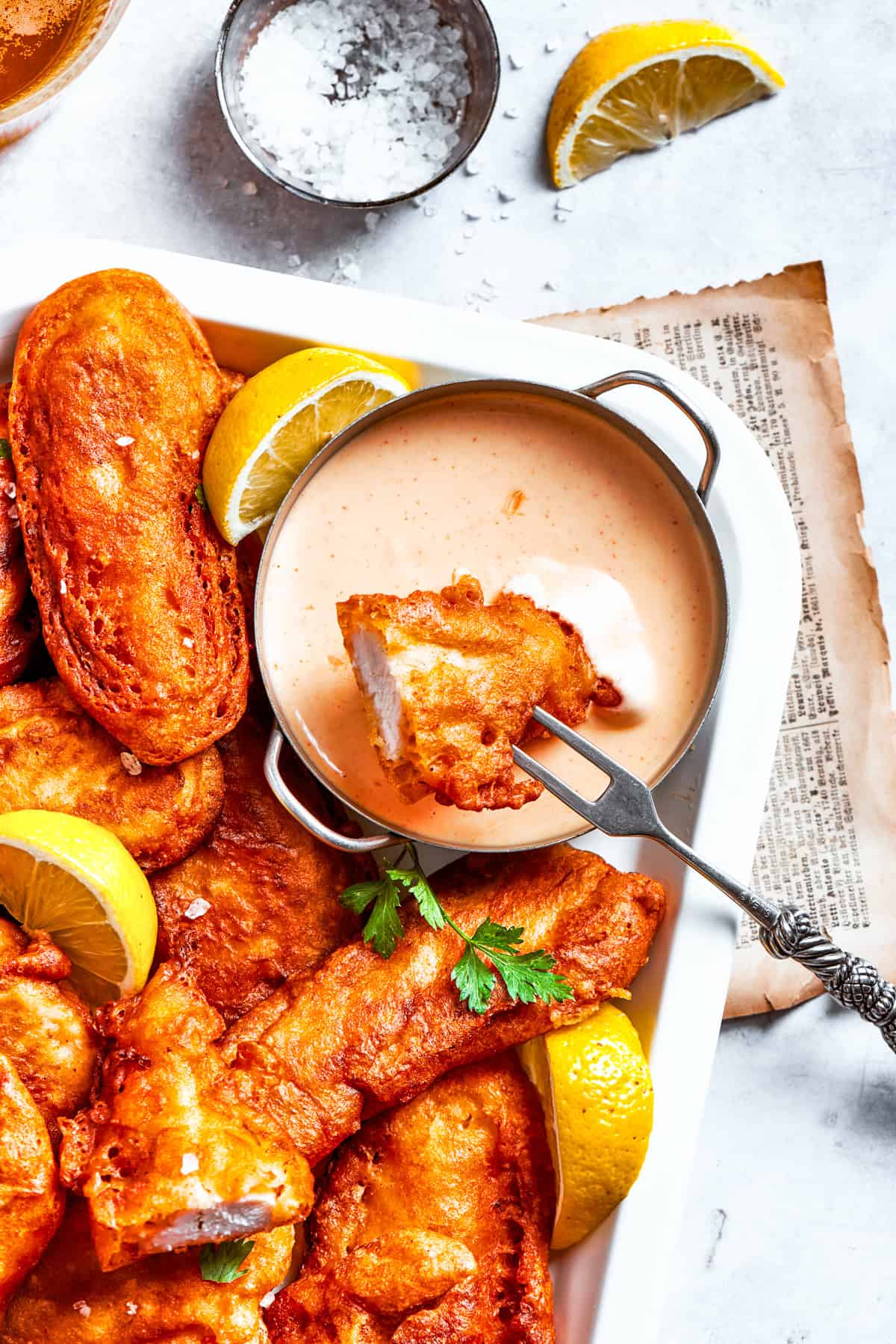 A piece of beer battered chicken is dipped into spicy sauce with more chicken pieces around it.