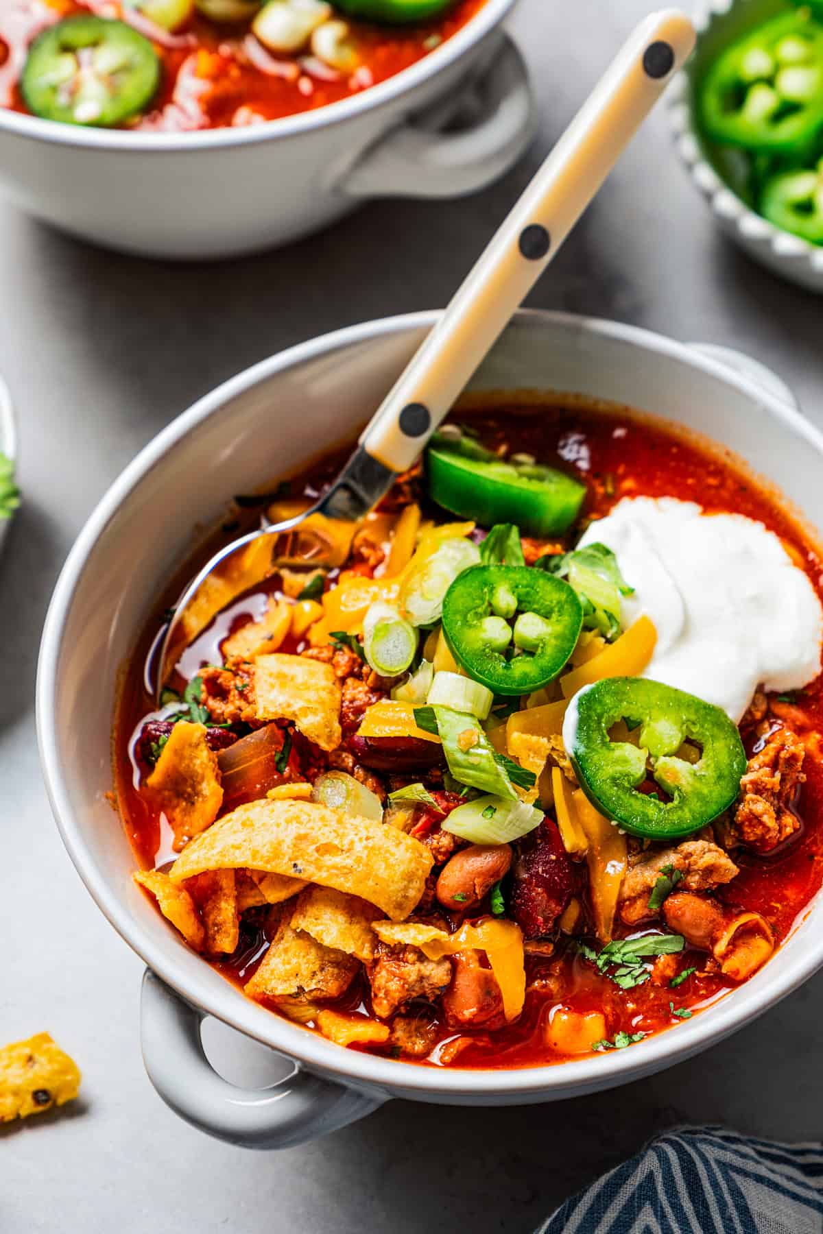 A bowl of turkey chili garnished with sour cream, jalapeños, and corn chips, with a second bowl in the background.