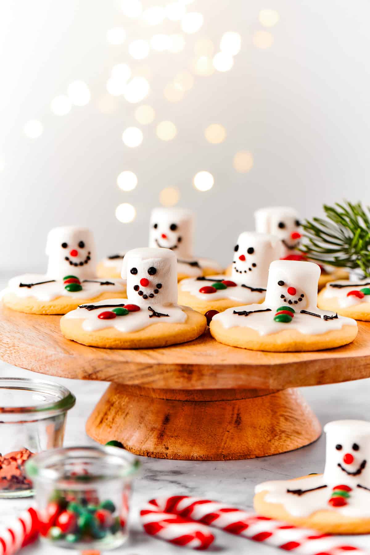 Melting snowman cookies are arranged on a wooden cake plate, with holiday decorations nearby.