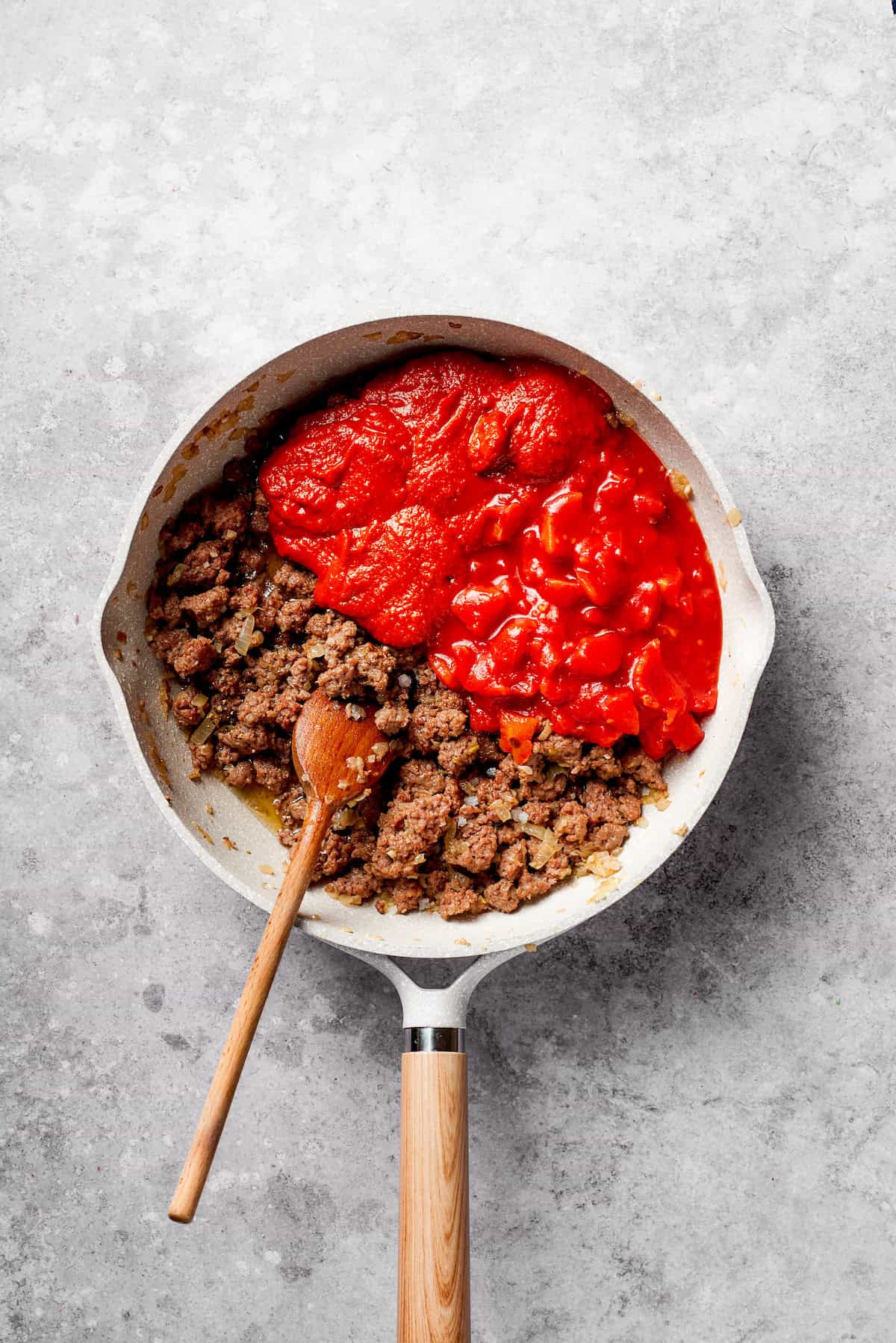 Canned tomatoes are mixed into ground beef in a frying pan.