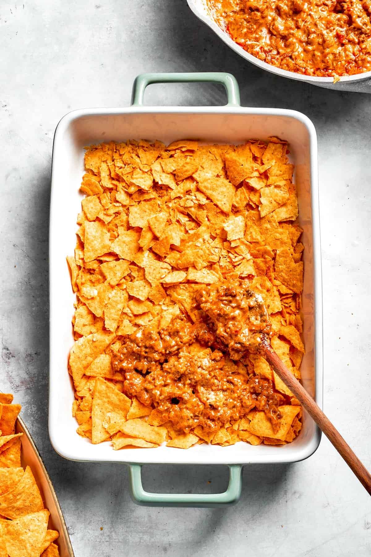 A casserole dish with a layer of Doritos being topped with beef filling.