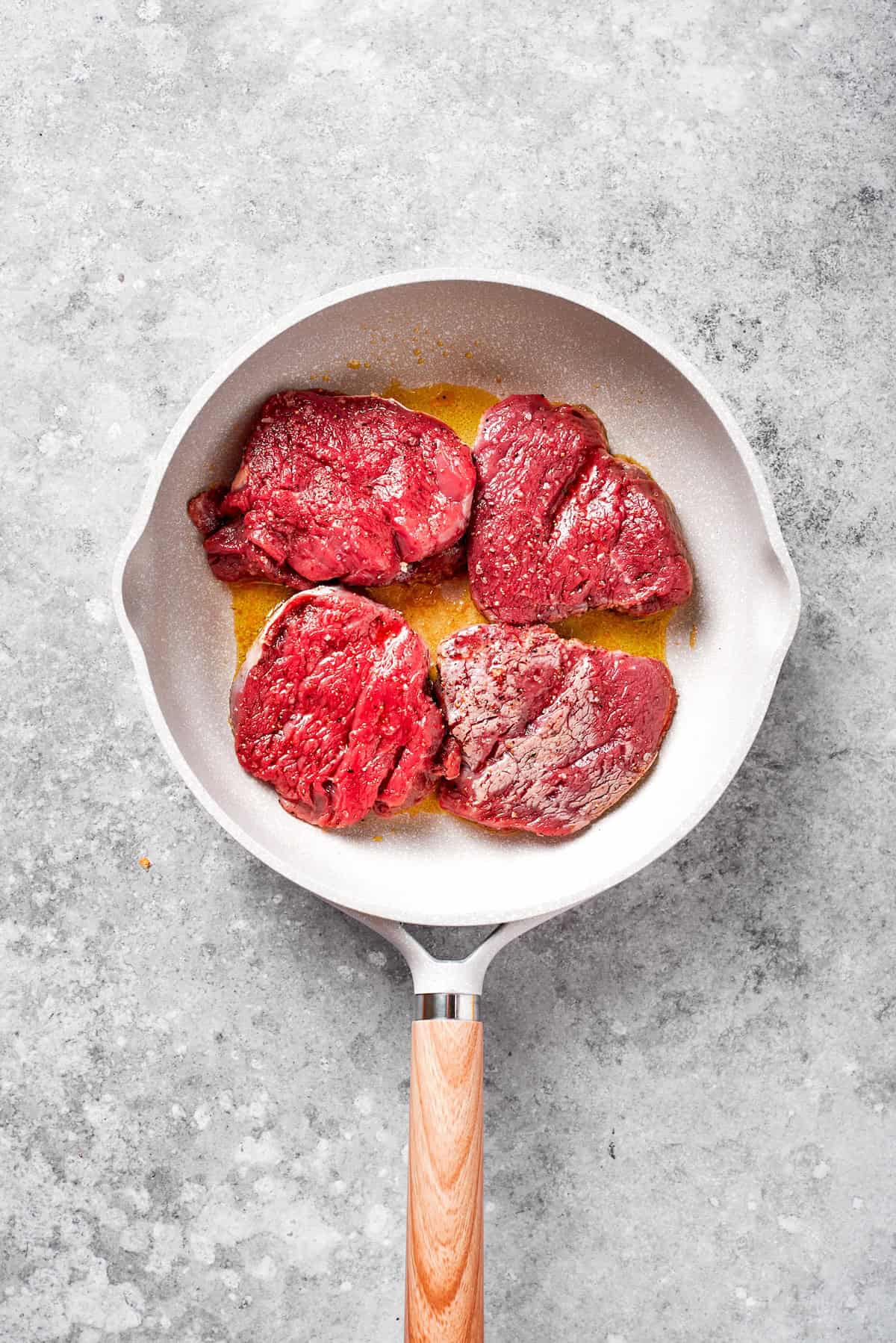 Four pieces of steak cook in a skillet.