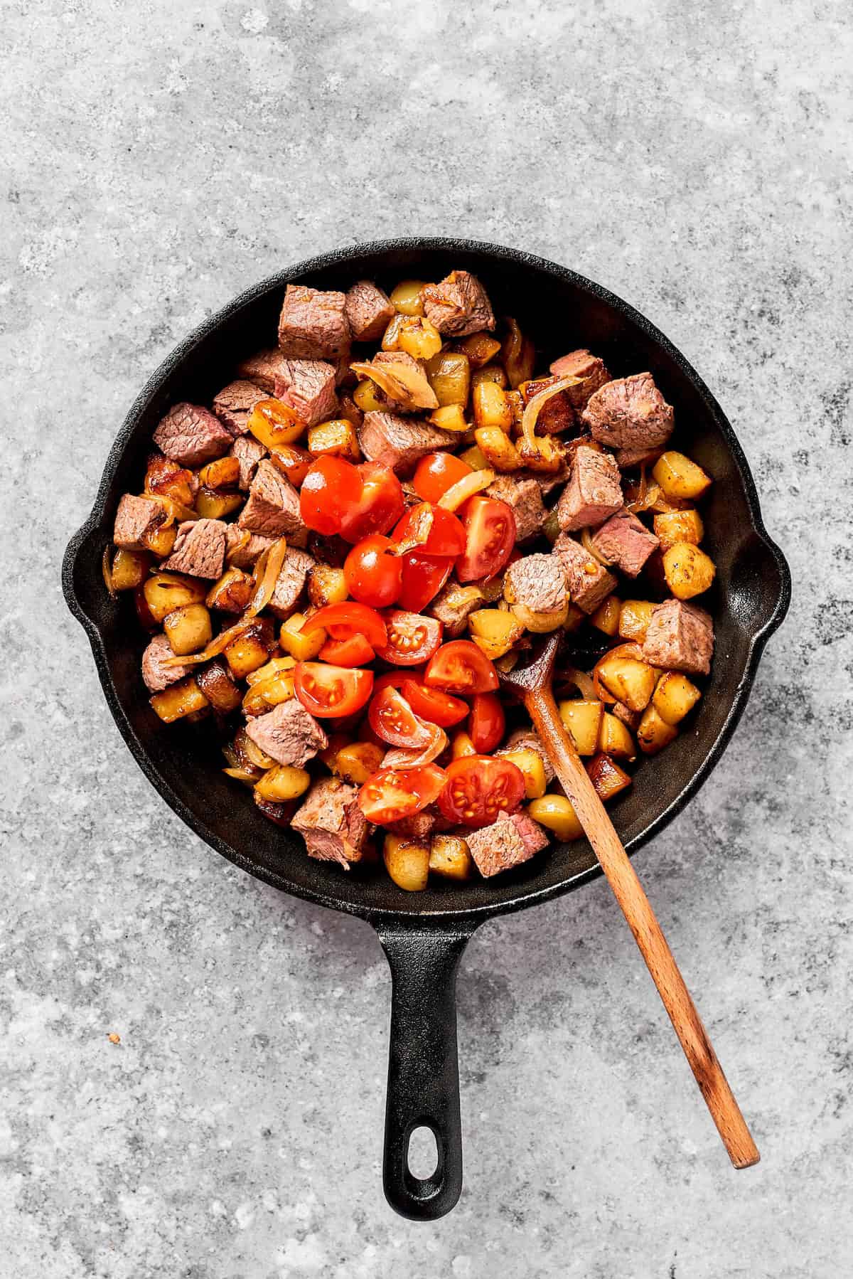 A spoon stirs a skillet of steak and potatoes.