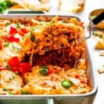 A serving spoon lifting out a slice of Dorito casserole from a baking dish.