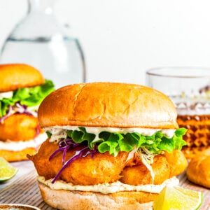 A fish sandwich on toasted buns with another sandwich and drinks placed behind it.