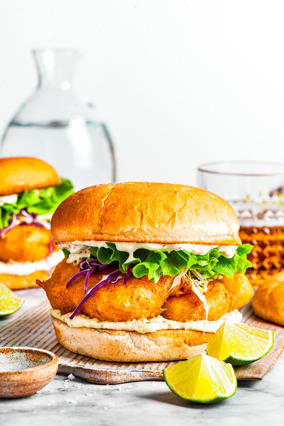 A fish sandwich on toasted buns with another sandwich and drinks placed behind it.