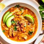 Spicy chicken soup in a bowl garnished with avocado slices and lemon wedges.