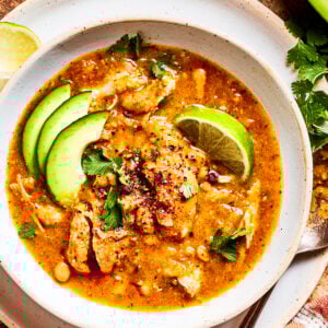 Spicy chicken soup in a bowl garnished with avocado slices and lemon wedges.
