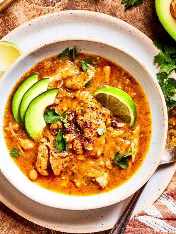 Spicy chicken soup in a bowl garnished with avocado slices and lemon wedges.