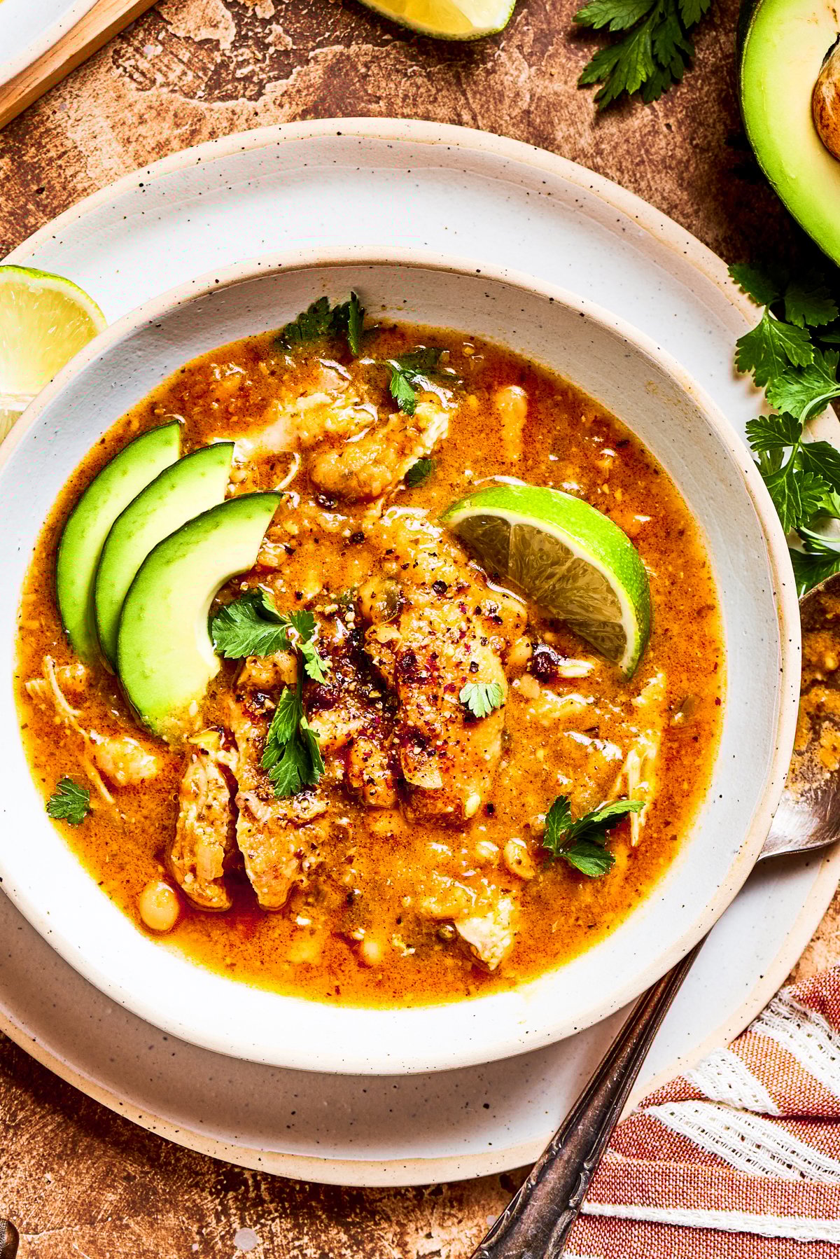 Spicy chicken soup in a bowl garnished with avocado slices and lemon wedges.